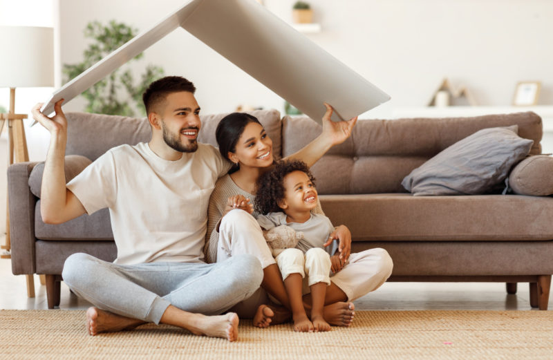 Cheerful familymultiethnic parents with child smiling and keeping roof mockup over heads while sitting on floor in cozy living room during relocation