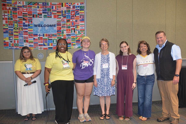 Seven people in front of a welcome sign with flags from all over the world