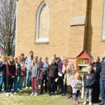 Group of people around a Little Library full of books in front of a church building with stained glass windows.