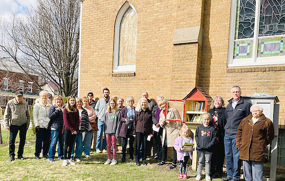 Group of people around a Little Library full of books in front of a church building with stained glass windows.