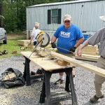 Men sawing a board in front of a mobile home.