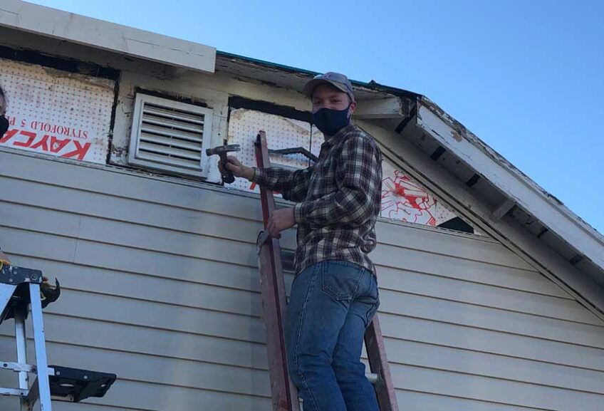 Man in hat, face covering, flannel shirt, and jeans hammering on a damaged house