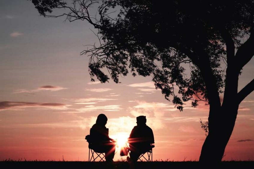 Two people sitting on chairs next to a tree, silhouetted against a pink sky and low sun