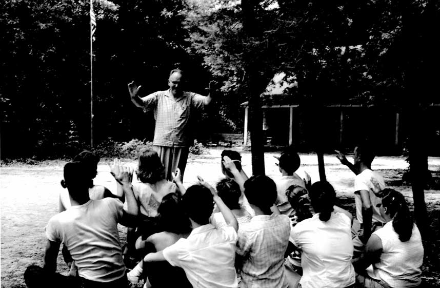 Black and white photo of Perry Huffaker conducting music with youth sitting on the ground near trees