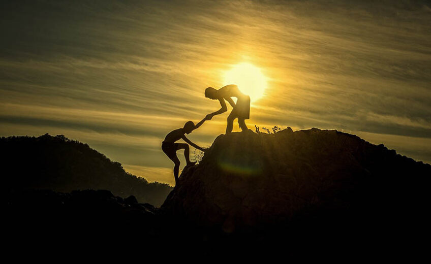 Person with the sun behind them helping another person up a steep hill