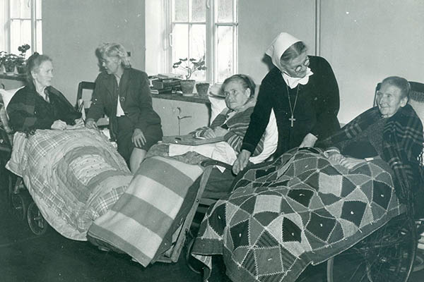 People in wheelchairs with handmade quilts. A woman with a white scarf on her head is leaning over one.