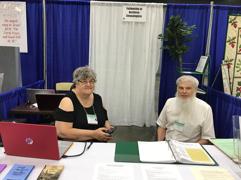 Two people sitting at a table in an exhibit hall booth with documents in front of them.