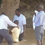 Four men stand in a river. One is kneeling to be baptized.