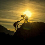 Person with the sun behind them helping another person up a steep hill